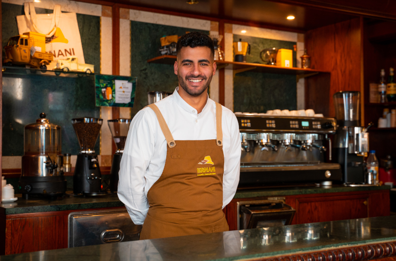 Amine, barista della Torrefazione Ernani a Milano, dietro al banco con la divisa: camicia bianca e grembiule marrone Ernani