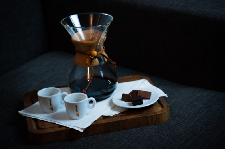 Foto stile Lifestyle con un vassoio rettangolare in legno scuro, con un tovagliolo bianco steso sopra e con appoggiato un piattino con dei cioccolatini, un fiore, due tazzine Ernani e la Chemex con il caffè filtro al suo interno, su sfondo grigio scuro.