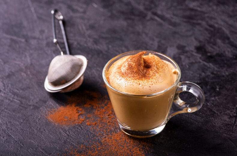 Foto di un tavolo in legno scuro con un filtro in metallo per il tè ed una tazzina in vetro piena di crema fredda al caffè, con una spolverata di cacao