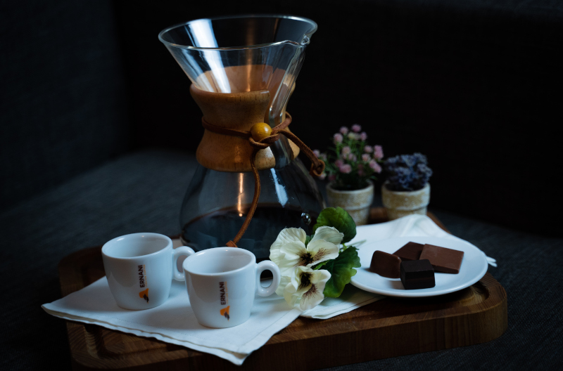 Foto con luci soffuse e atmosfera rilassata con un vassoio in legno sopra al quale appoggiano due tazzine da caffè ernani, un piccolo mazzo di fiori, dei cioccolatini e la protagonista Chemex con il caffè all'americano al suo interno.