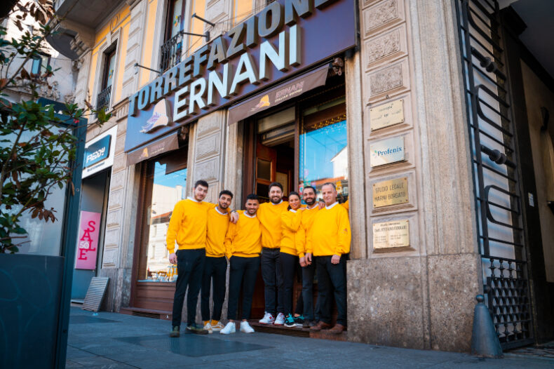 Foto della vetrina della Torrefazione con tutto il team di Caffè Ernani con la divisa (felpa gialla e pantaloni neri) che si abbracciano e sorridono.