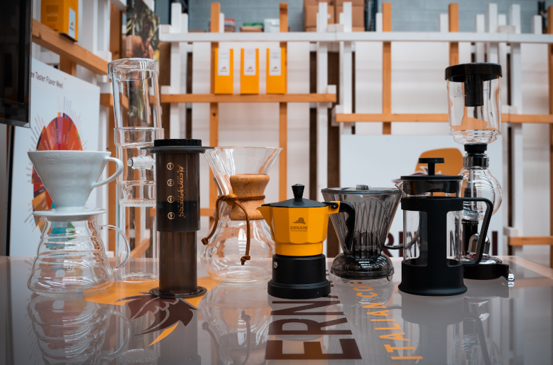 Foto del tavolo Ernani con sopra tutti gli strumenti a disposizione per poter preparare il caffè filtro o il caffè all'americana. Partendo da sinistra: V60, Cold brew Dripper, Aeropress, Chemex, Moka, Clever, French press, Syphon.