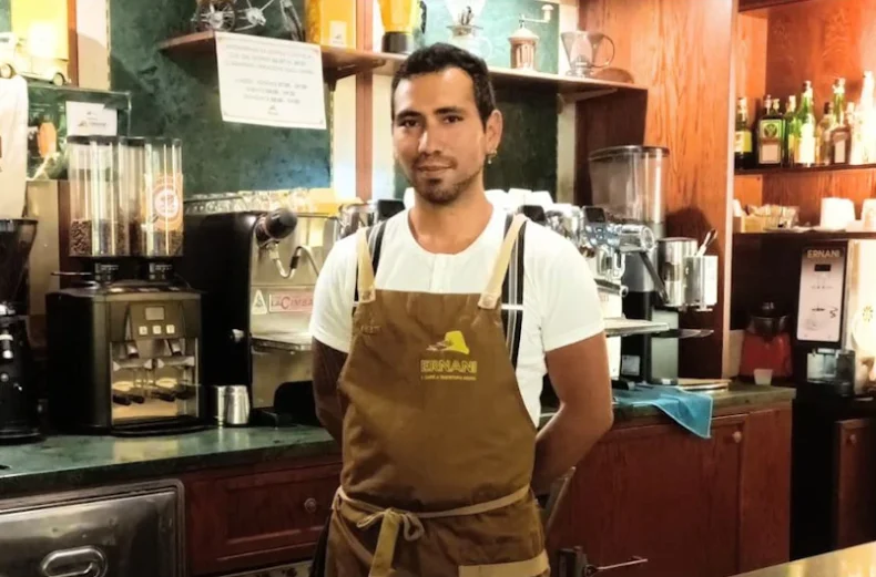 Foto di Cristian, un barista della Torrefazione Ernani a Milano con la divisa estiva (grembiule Ernani e maglietta bianca mezze maniche) dietro al bancone del locale.