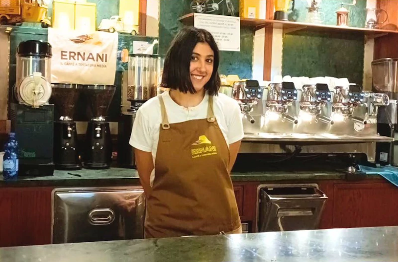 Foto di Francesca, una barista della Torrefazione Ernani a Milano con la divisa estiva (grembiule Ernani e maglietta bianca mezze maniche) dietro al bancone del locale.