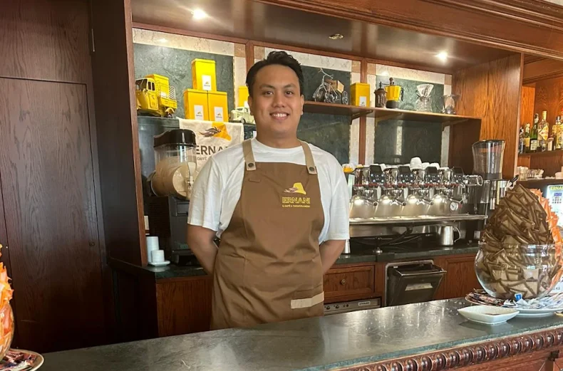 Foto di Mark, un barista della Torrefazione Ernani a Milano con la divisa estiva (grembiule Ernani e maglietta bianca mezze maniche) dietro al bancone del locale.