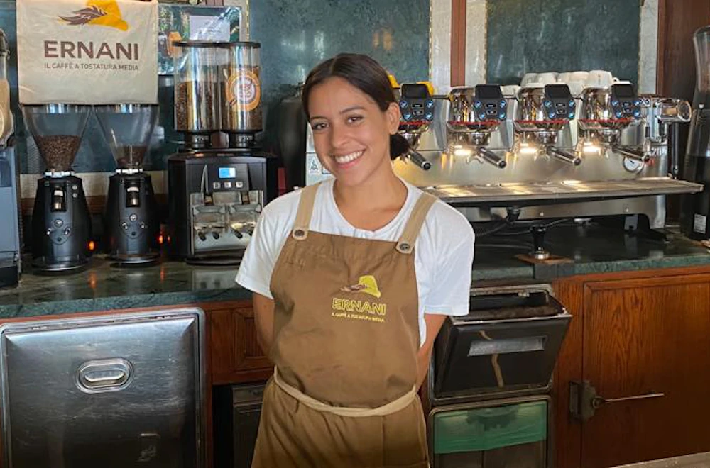 Foto di Stephannie, una barista della Torrefazione Ernani a Milano con la divisa estiva (grembiule Ernani e maglietta bianca mezze maniche) dietro al bancone del locale.